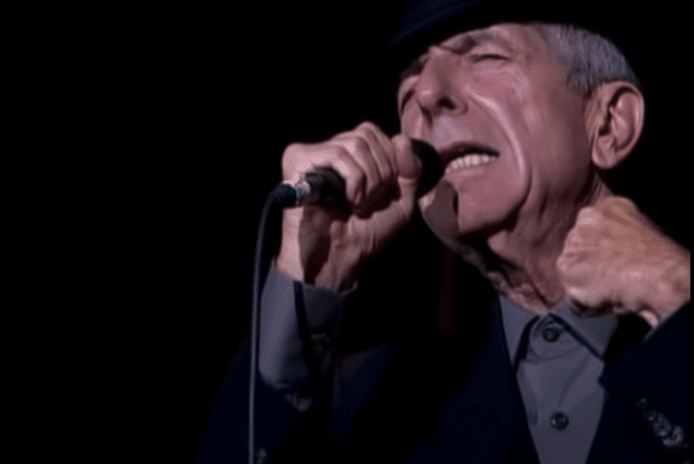 Leonard Cohen, eyes closed and fist clenched, singing and illuminated by a spotlight.