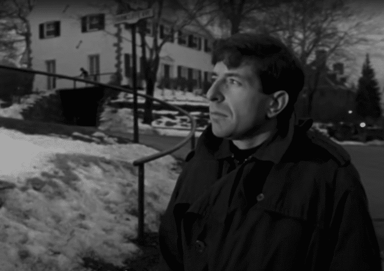 A young Leonard Cohen walks in front of a large house, gazed intently at something out of the frame. The ground is covered in snow.