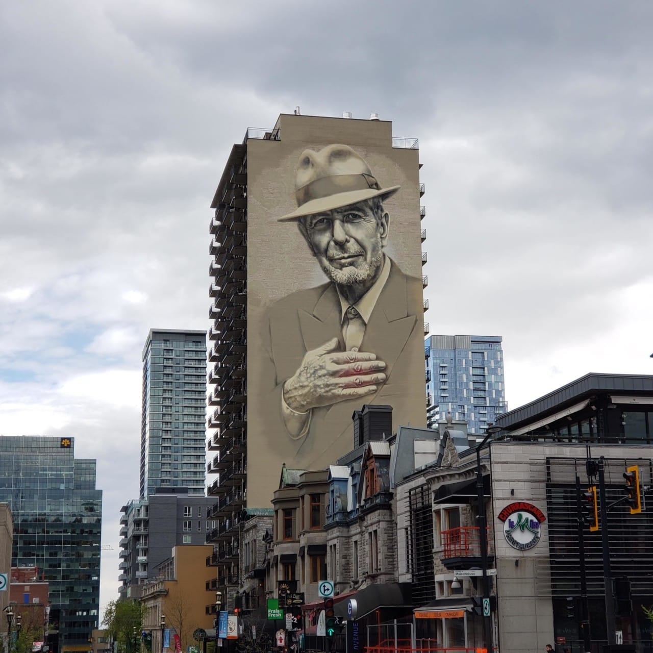 A tall apartment building looms over typical Montreal row houses, with a 12-story mural of Leonard Cohen, hand placed on his chest, fingers parallel to the ground. A red glow is visible behind his fingers.