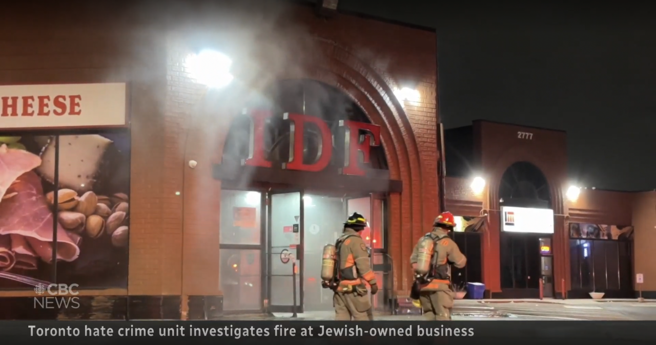 A photo of a grocery store with a sign reading “IDF”, with smoke coming out the front doors, and two firefighters in front of it.